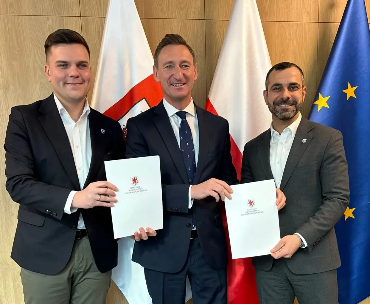 Sygnatariusze umowy prezentują podpisany dokument, w tle flagi.