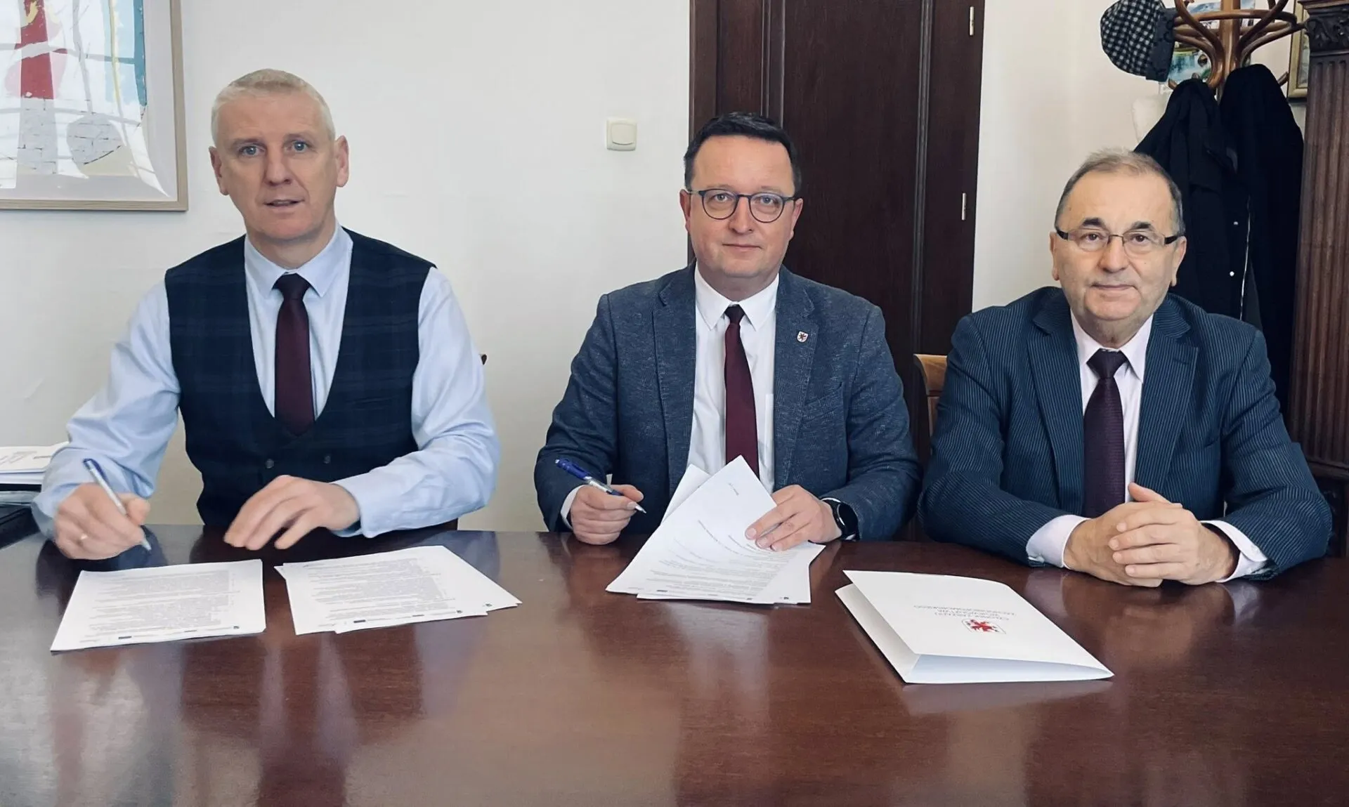 Strony porozumienia podpisują dokument przy stole prezydialnym, w tle biała ściana i drzwi.