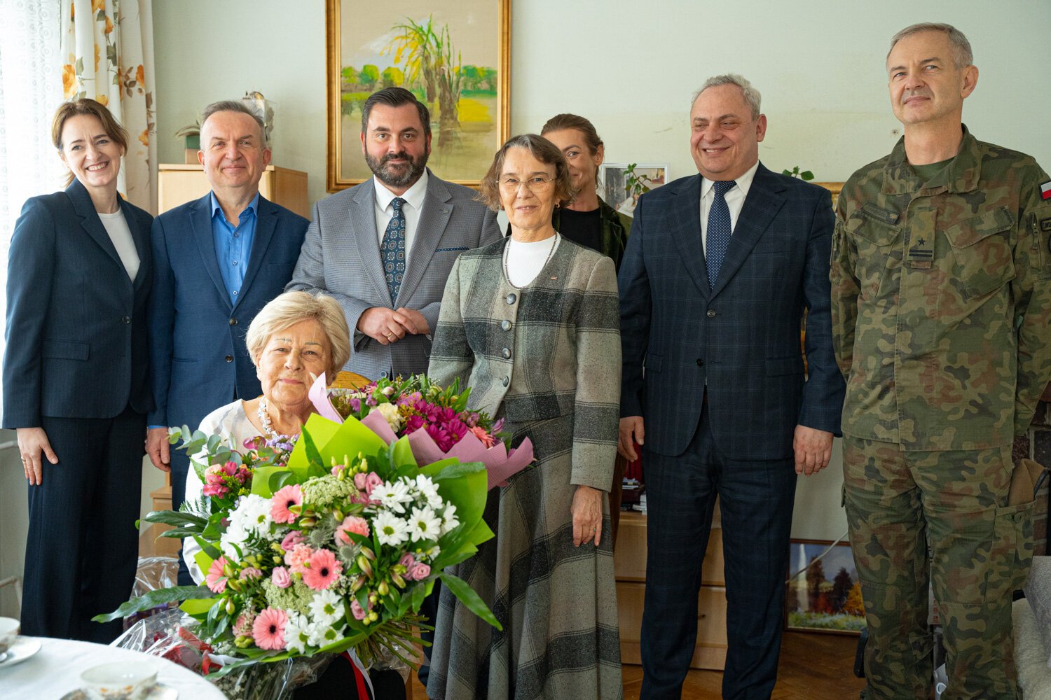 Goście uroczystości pozują do wspólnego zdjęcia, przed nimi jubilatka z bukietami kwiatów oraz stół z upominkami.
