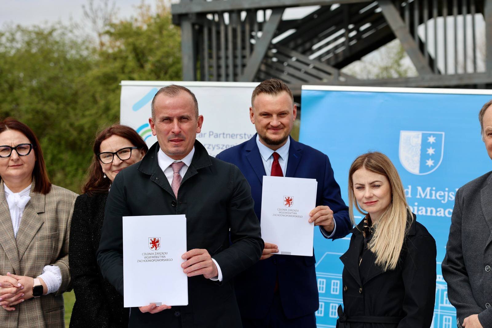 Strony umowy prezentują podpisane porozumienie, w tle ścianki promocyjne oraz infrastruktura turystyczna.