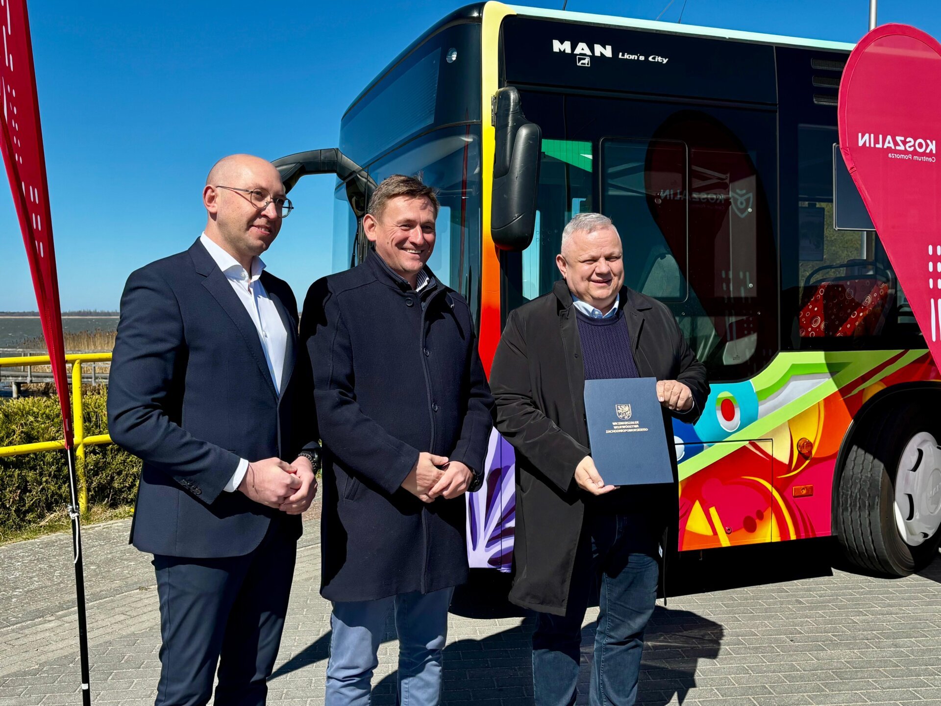 Strony porozumienia prezentują przekazaną umowę, w tle jezioro i autobus MZK Koszalin.