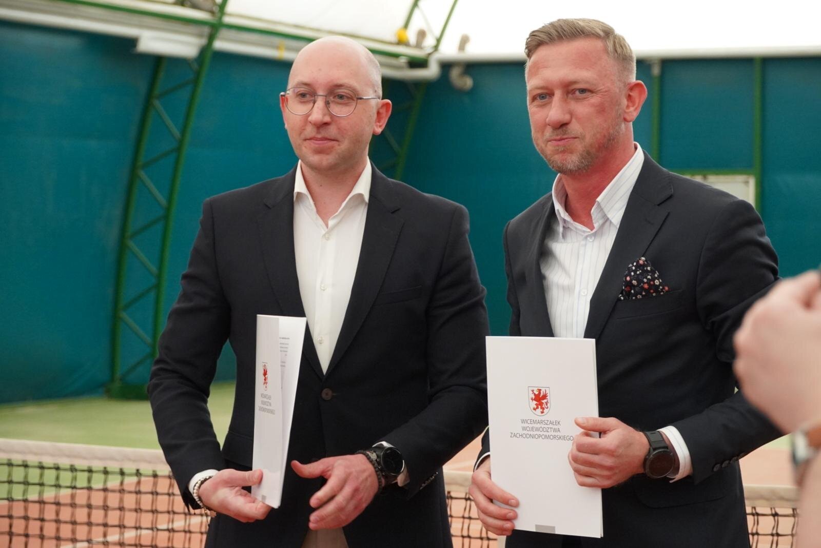 Strony umowa prezentują podpisane porozumienie, w tle kort tenisowy.