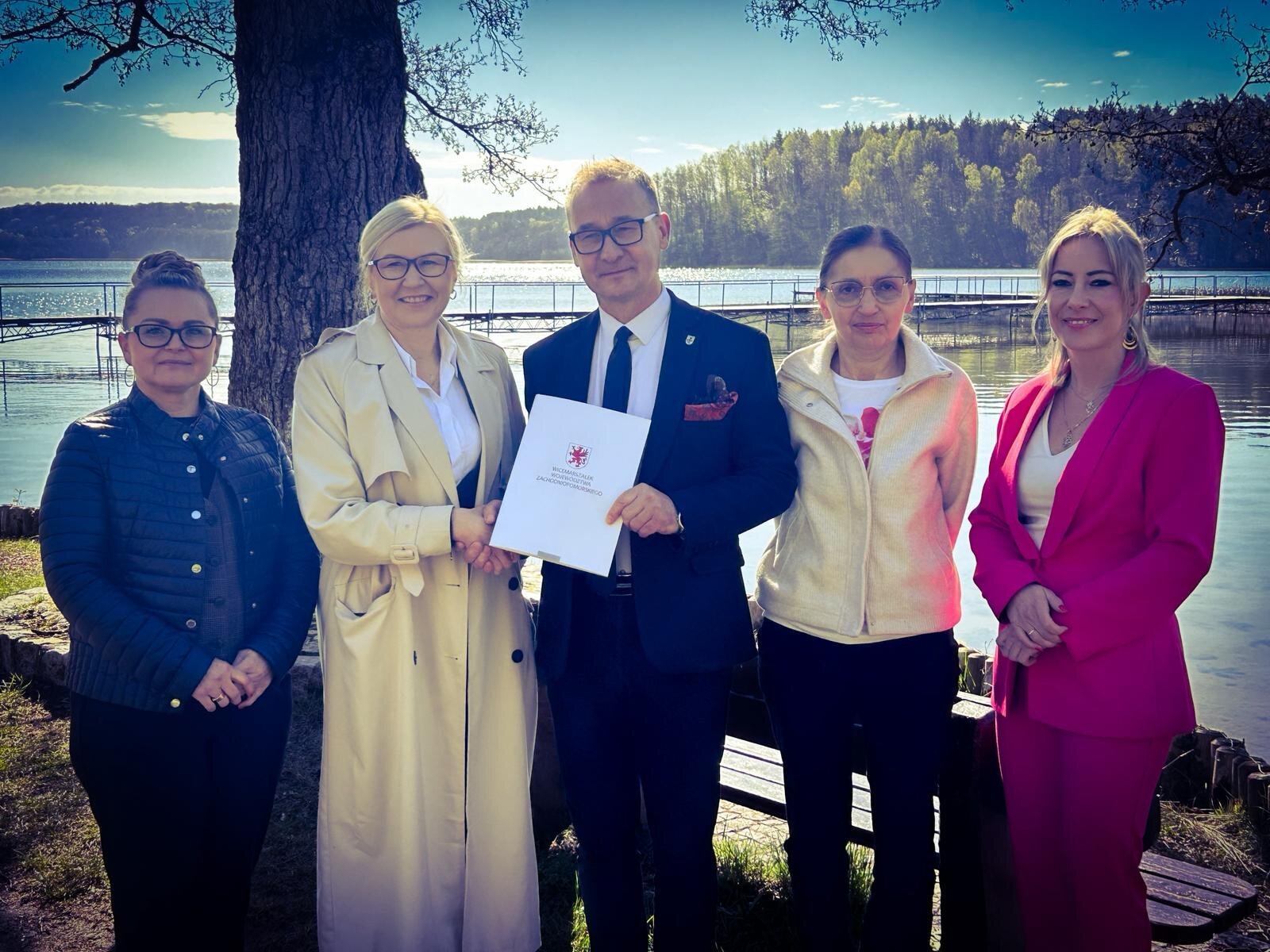 Strony umowy prezentują podpisany dokument, w tle plaża i jezioro.