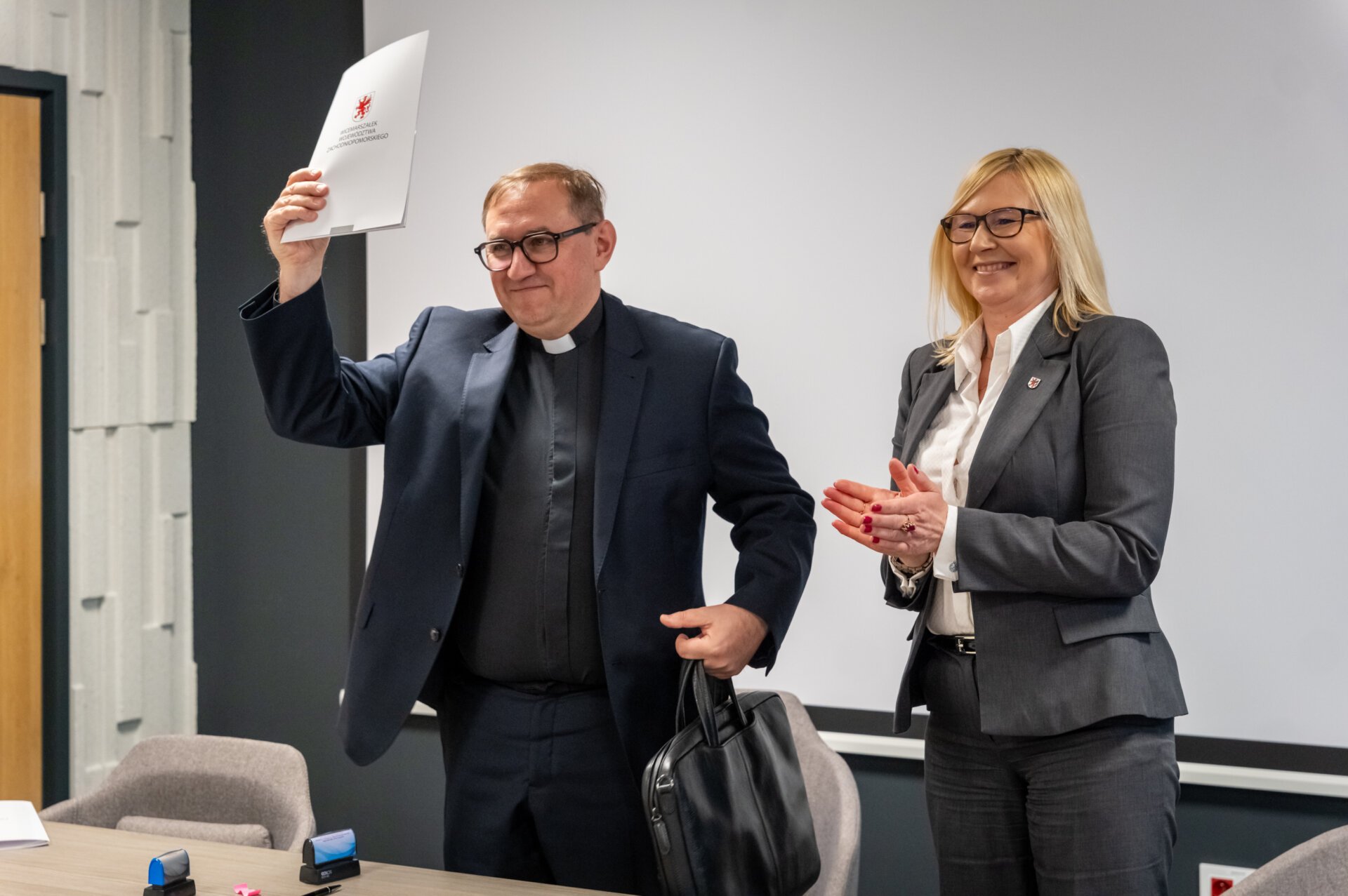 Ksiądz podnosi teczkę z umową, obok stoi wicemarszałek Anna Bańkowska