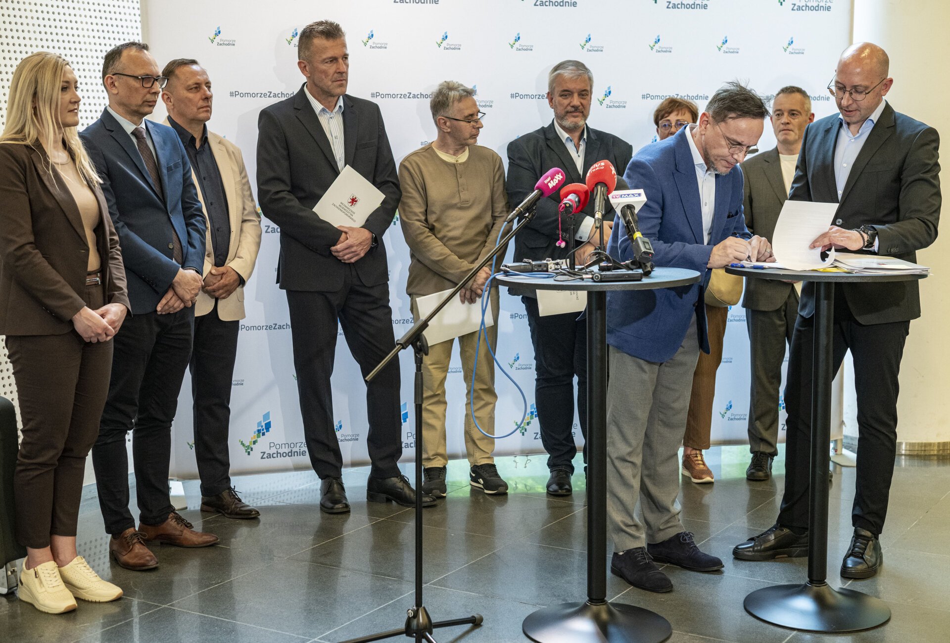 podpisanie umów podczas konferencji prasowej, część gości stoi przy stolikach koktajlowych, na stolikach są dokumenty i mikrofony mediów, w tle pozostali goście