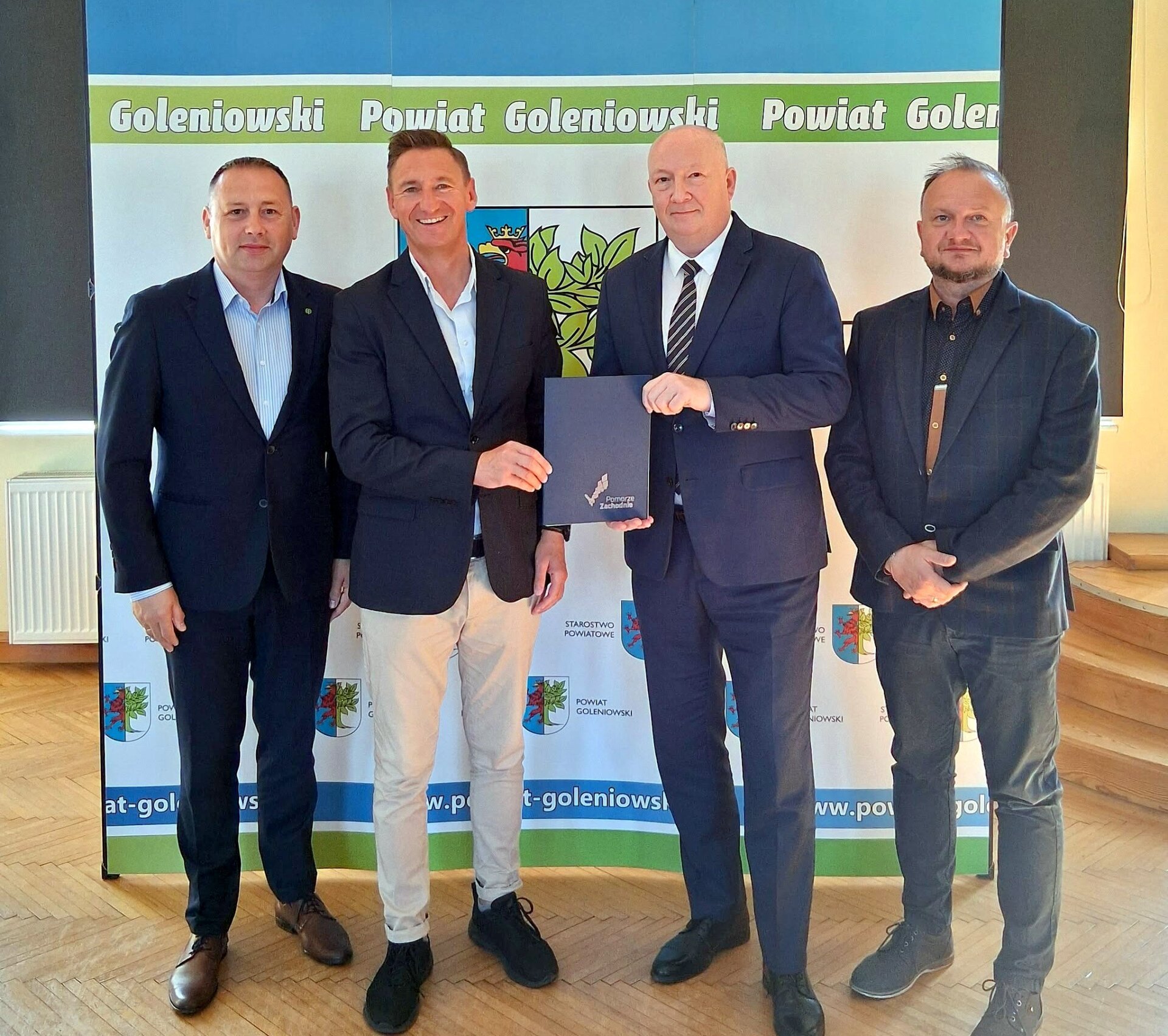 Strony umowy prezentują podpisany dokument, w tle ścianka promocyjna Powiatu Goleniowskiego - fot. Starostwo Powiatowe w Goleniowie.
