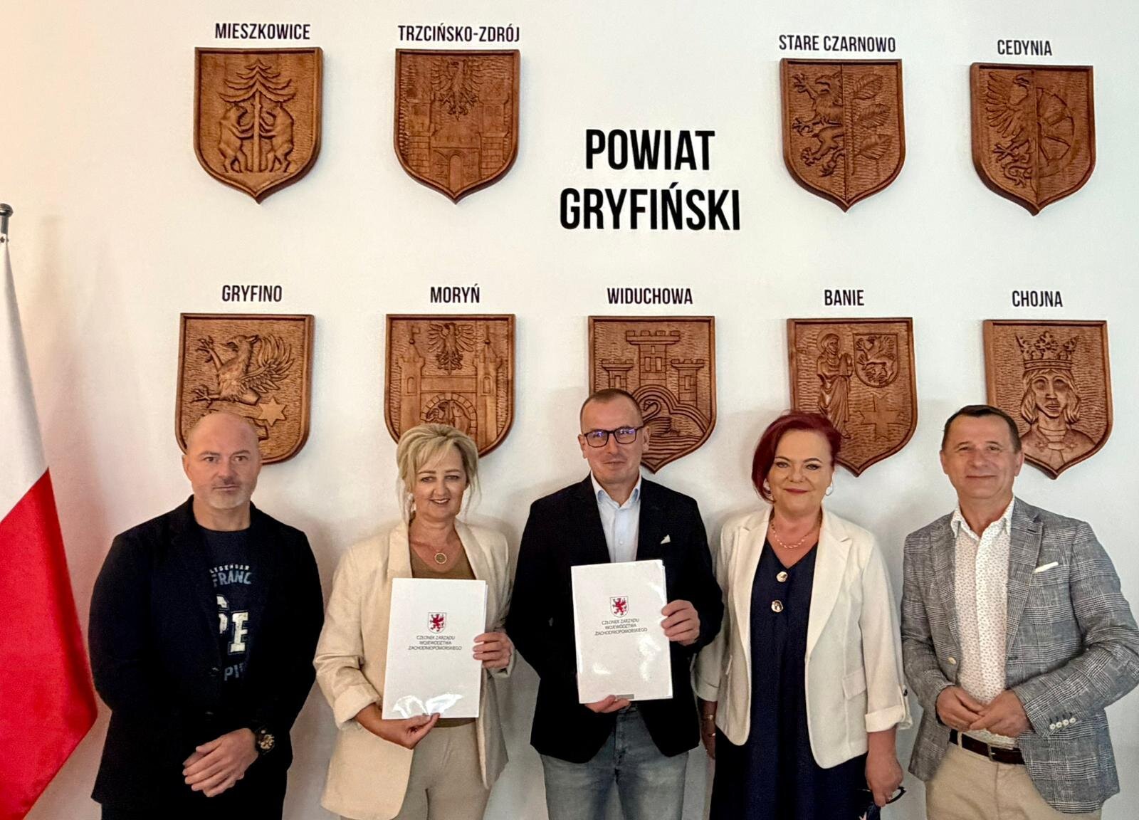 Strony umowy prezentują podpisany dokument, w tle ściana z herbami gmin tworzącymi Powiat Gryfiński.