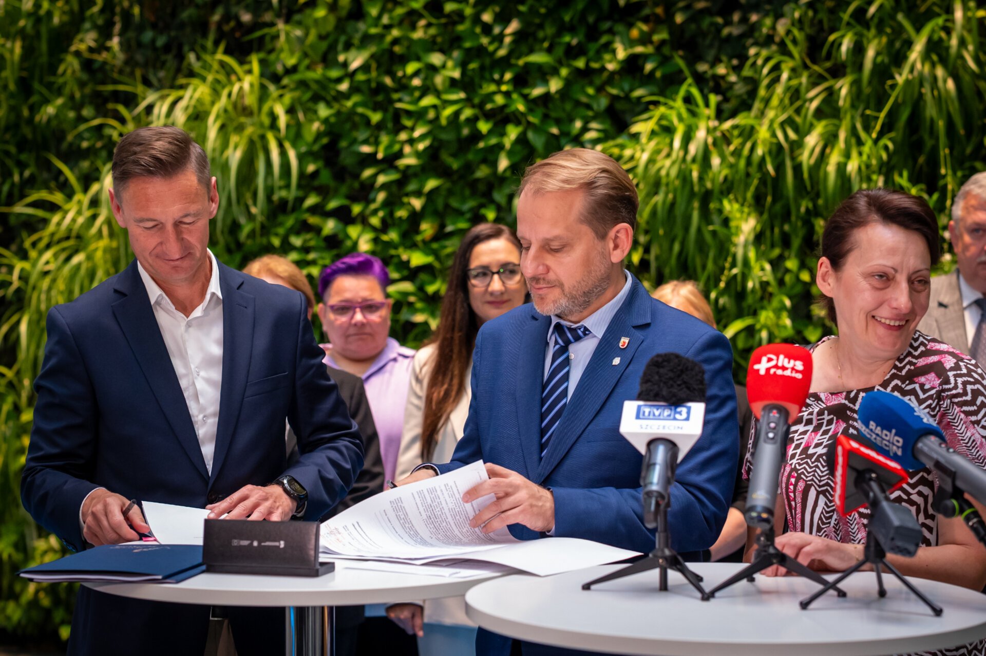 Strony umowy podpisują dokument przy stoliku, w tle goście spotkania i zielona ścianka w atrium w Urzędzie Marszałkowskim.