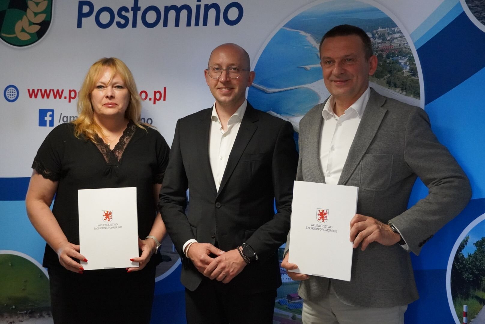 Strony umowy prezentują podpisany dokument, w tle ścianka promocyjna Gminy Postomino.