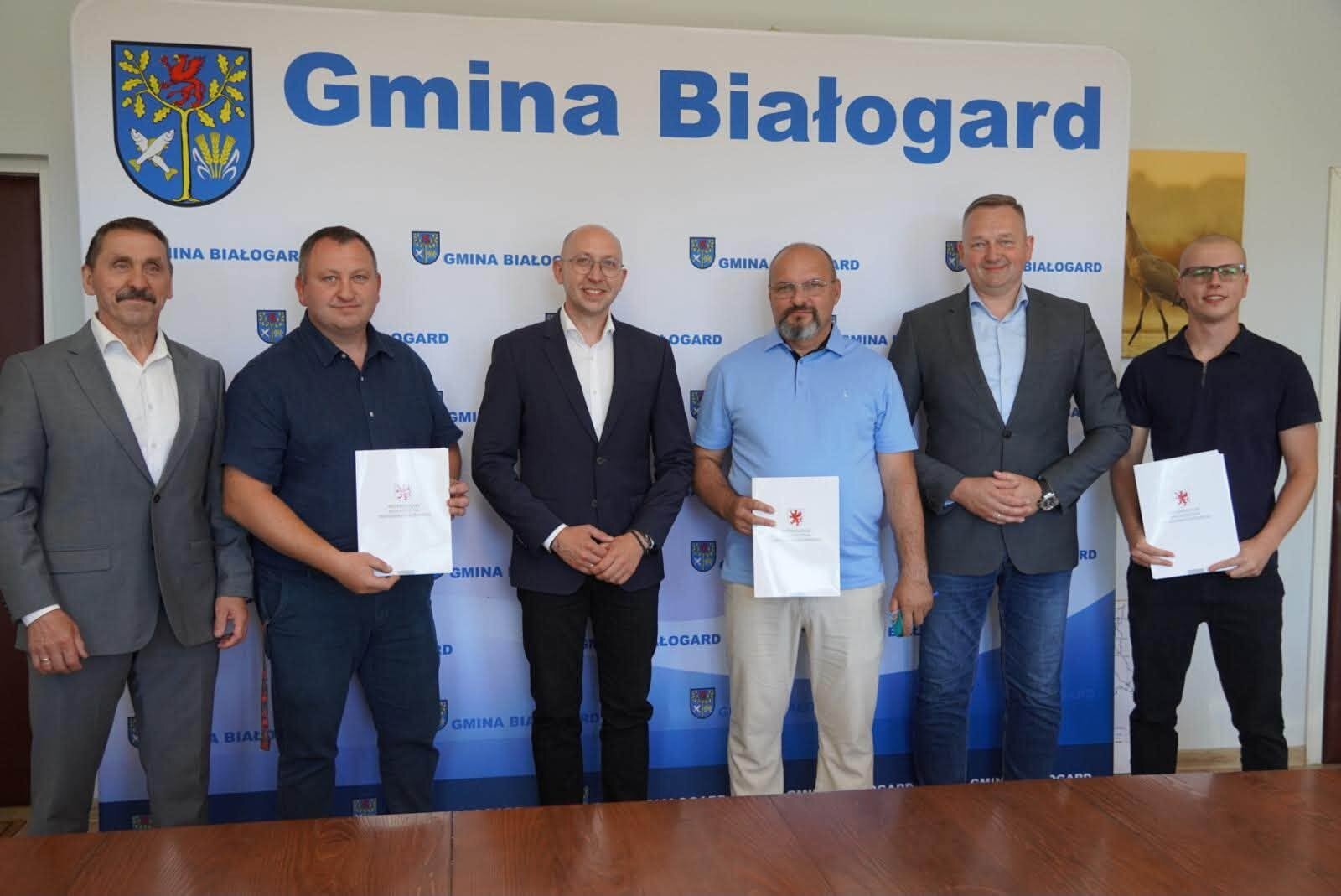 Sygnatariusze umów prezentują podpisane dokumenty, w tle ścianka promocyjna Gminy Białogard.