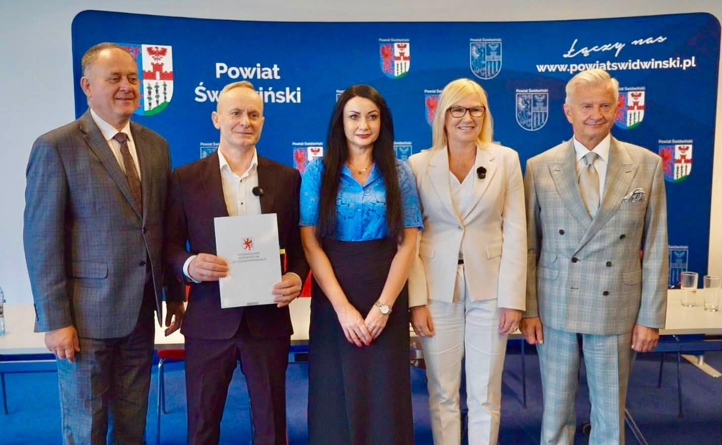 zdjęcie 5 osób, w wicemarszałek Bańkowskiej w jasnym garniturze z beneficjentami stoi na tle niebieskiej ścianki,