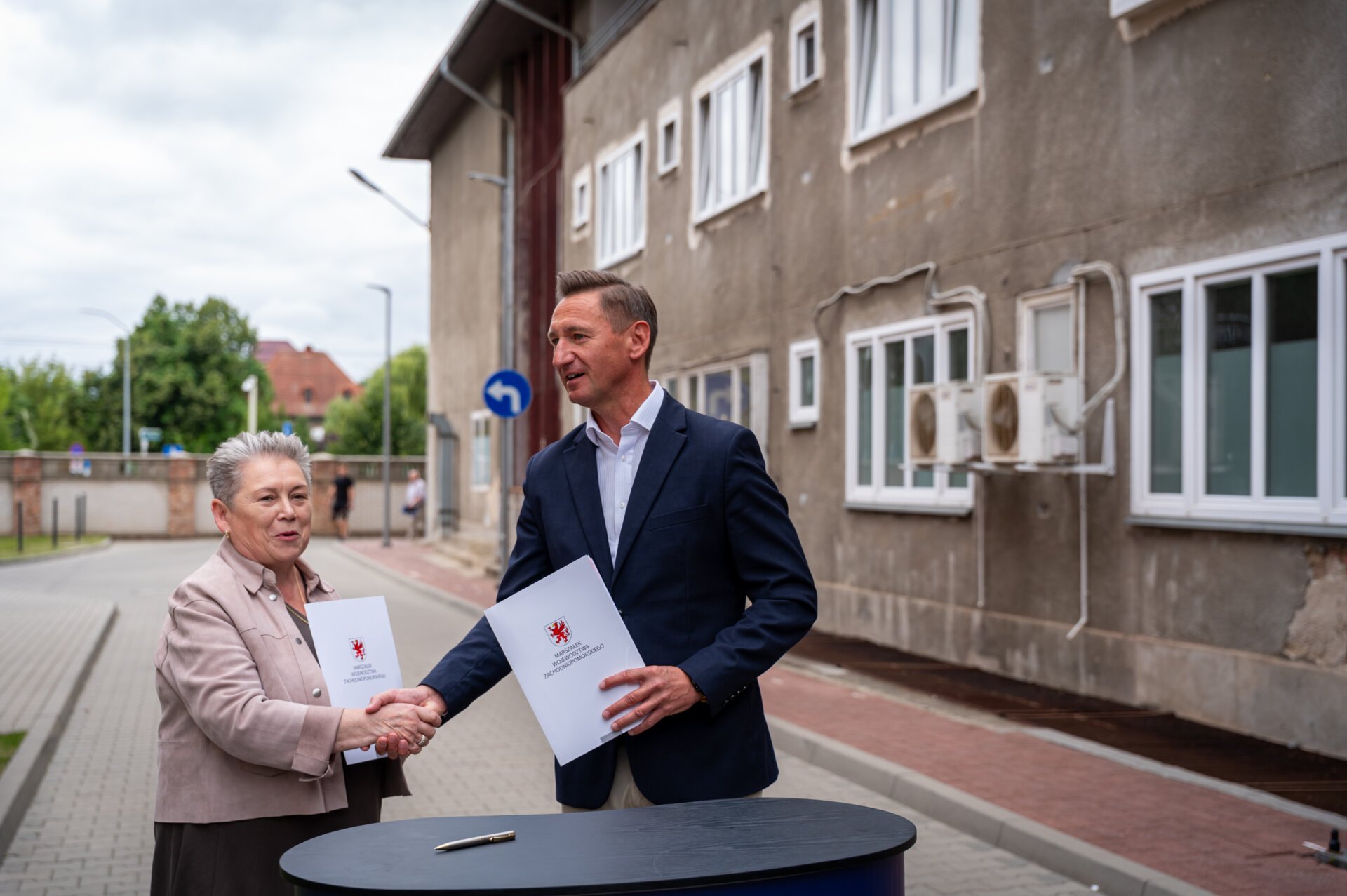 Strony umowy gratulują sobie podpisania porozumienia, w tle budynek przewidziany do remontu.