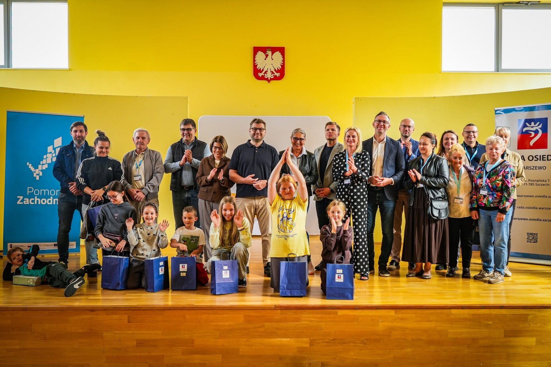 zdjęcie na scenie na sali gimnastycznej uczestników festynu osiedlowego na Warszewie, na zdjęciu młodzież, kobiety i mężczyźni, wszyscy uśmiechnięci.