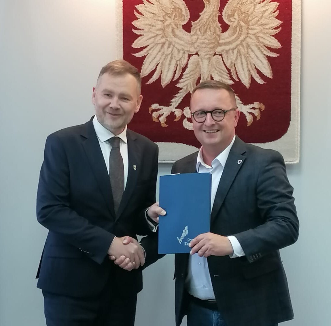 Strony umowy prezentują podpisany dokument, w tle godło Polski.